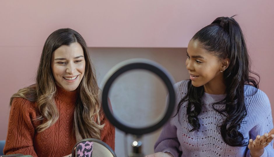 Diverse friends talking about makeup and smiling while having online meeting near ring lamp in beauty salon