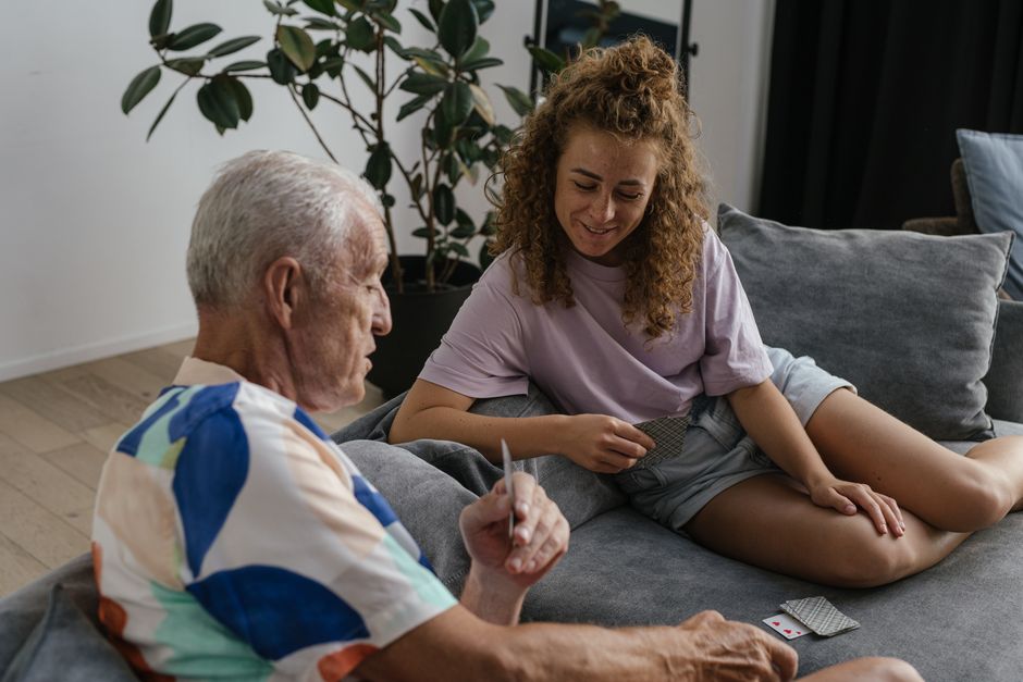 A senior adult and young woman share a joyful moment playing cards indoors on a cozy couch.