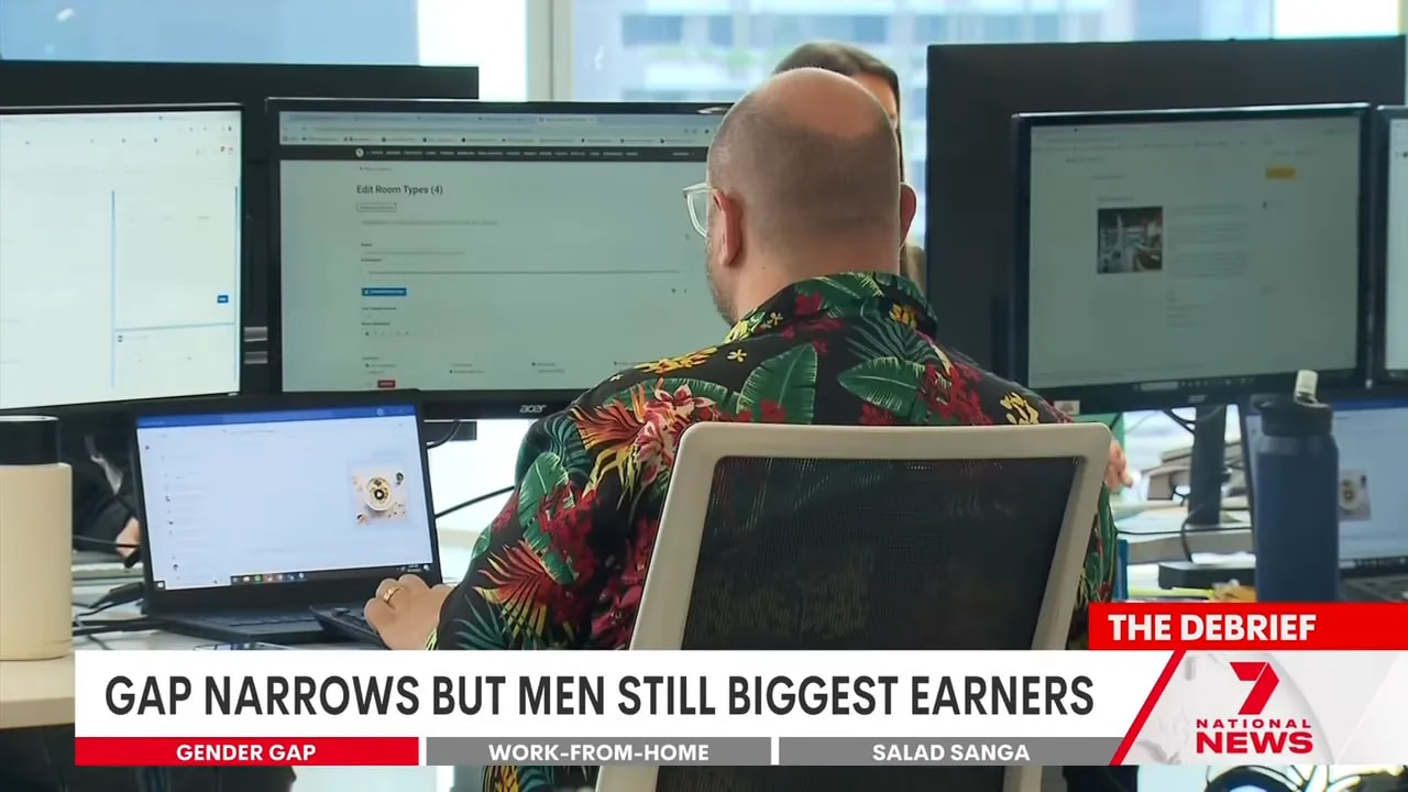 Rear view of an office worker in a floral shirt using a laptop and several monitors in an open-plan office.