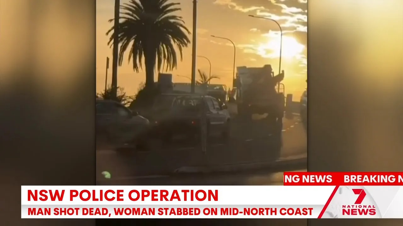 Sunset view of cars and a truck on a coastal road with on-screen text reading 'NSW Police Operation' and 'Man shot dead, woman stabbed'.