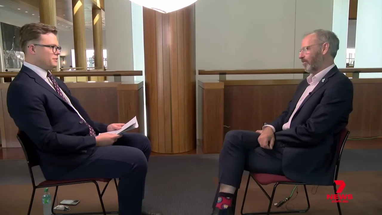 Full-frame wide shot of an interviewer and interviewee seated opposite each other in a formal indoor setting, providing context for a policy discussion; 7 News logo visible.