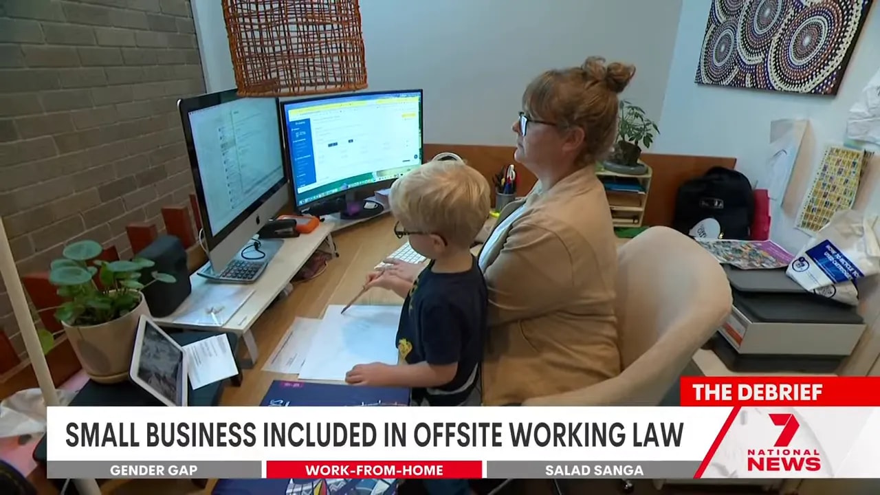 Home office scene showing an adult at a desk with dual monitors working while a young child stands beside them, illustrating practical realities of working from home.