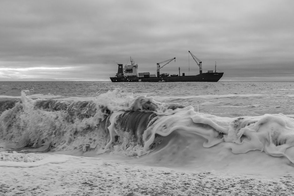 A cargo ship sails through rough ocean waves in a dramatic black and white scene.