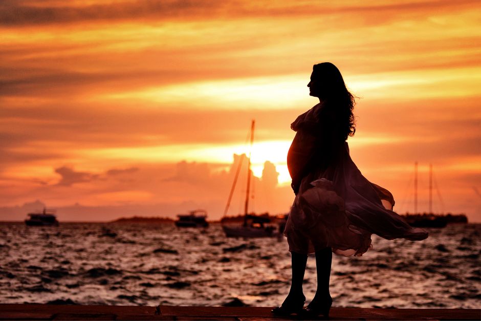 A serene silhouette of a pregnant woman standing by the sea during a tropical sunset.