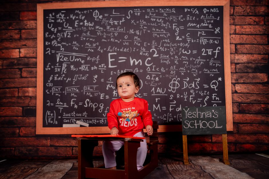 A baby in a classroom setting with a math-filled chalkboard background.