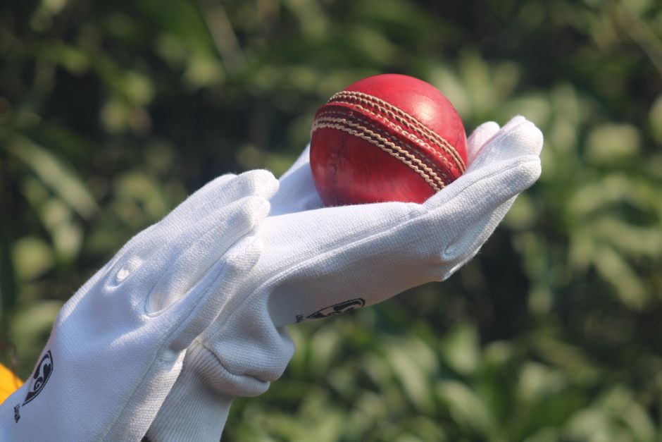 Detailed photo of a red cricket ball held by gloved hands outdoors.