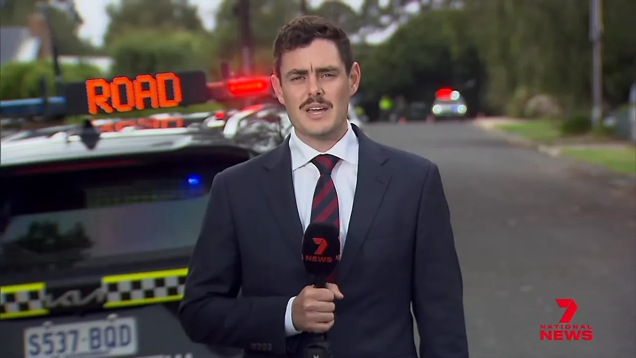 On‑scene news reporter holding a microphone with a police vehicle and flashing lights in the background on a suburban street.