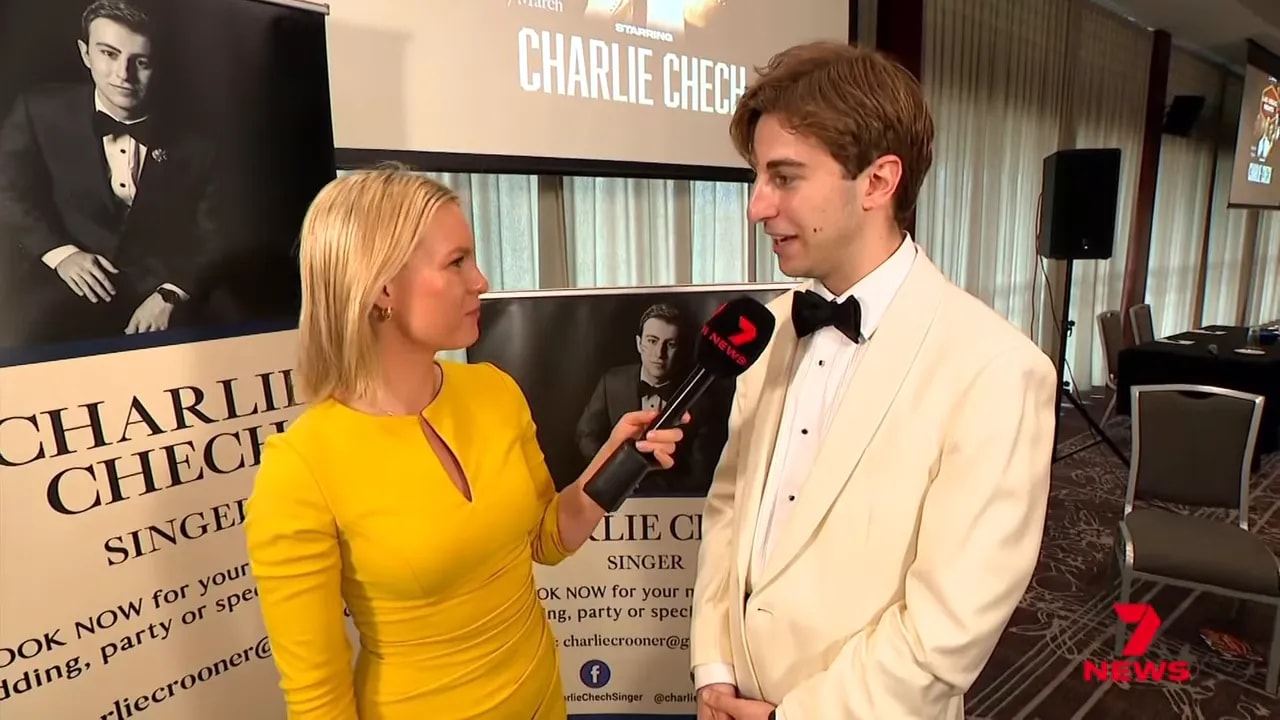Wide shot of a presenter in yellow interviewing a singer in a white jacket with promotional posters and a conference room visible