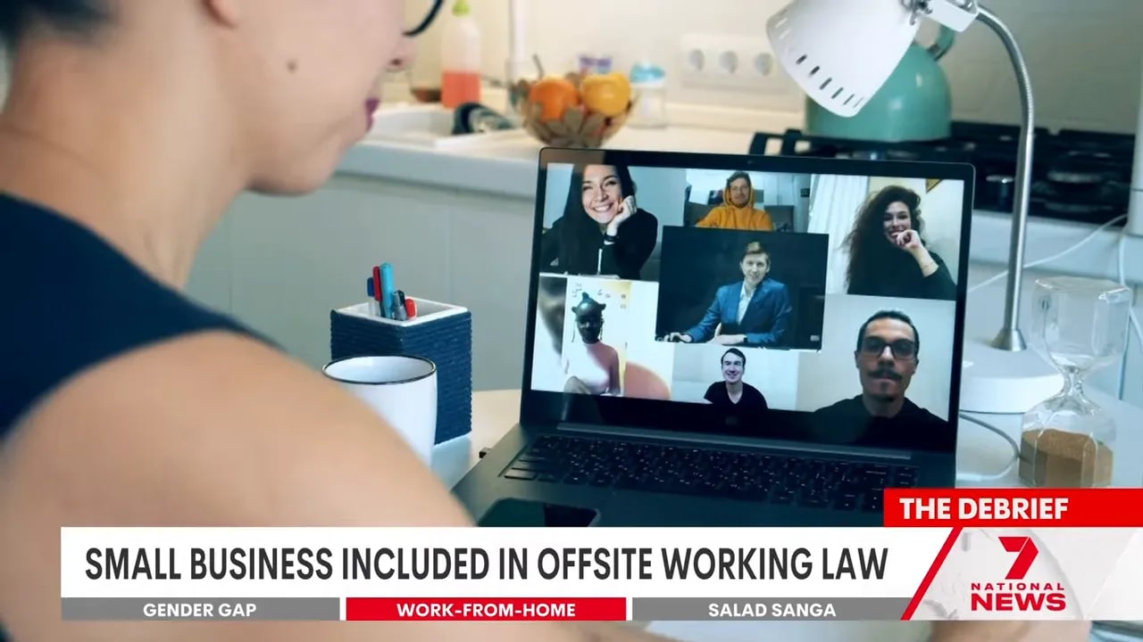 Person viewing a grid of colleagues in a video conference on a laptop at a home desk with desk lamp, mug and kitchen background.