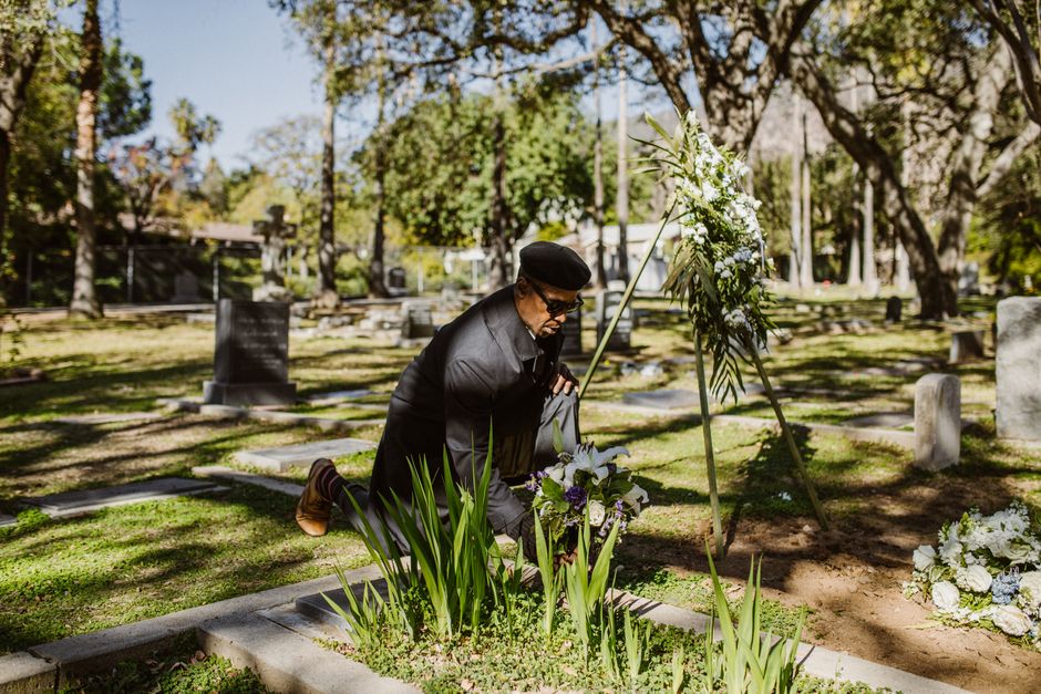 An elderly man in a cemetery mourning with flowers, surrounded by trees.