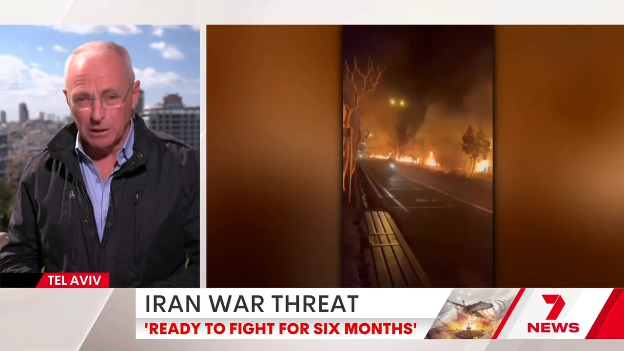 Vertical witness video shown on a news split-screen: a nighttime street with a bench and a long line of fires and smoke along the roadside.