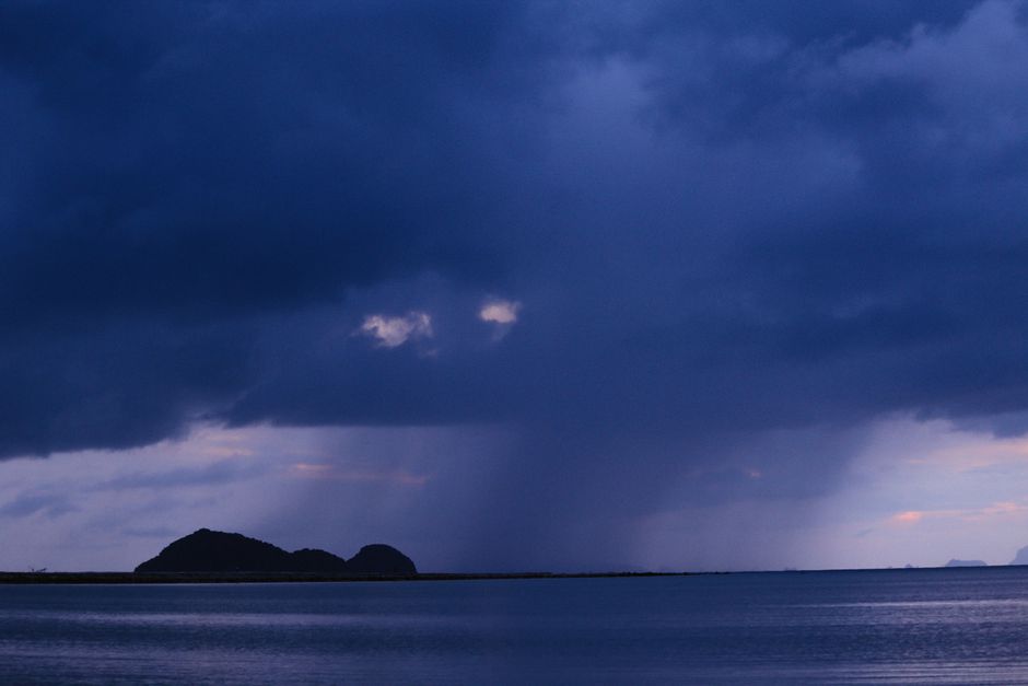 Silhouette of an island under dark rain clouds at twilight over the calm sea.